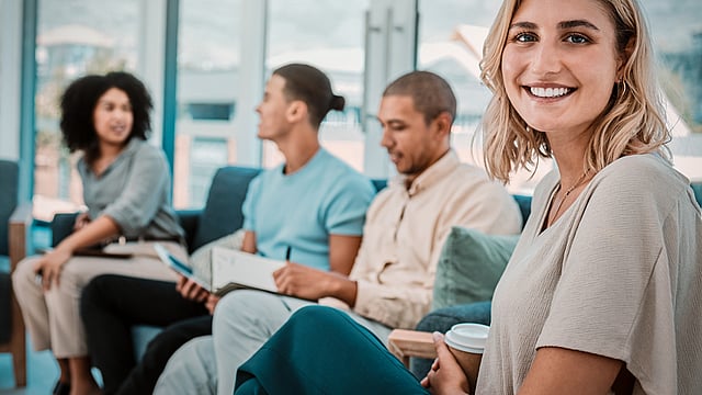 A woman is smiling and looking at the reader, with a group chatting in the background.